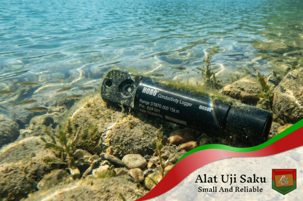 Gambar bawah air yang menunjukkan alat sensor level air dan konduktivitas merk HOBO, berbentuk silinder hitam, tergeletak di antara bebatuan dan pasir di dasar danau yang jernih. Perangkat tersebut sedikit tertutup ganggang dan dikelilingi oleh ikan-ikan kecil yang berenang serta tanaman air, dengan cahaya matahari menembus dari permukaan.